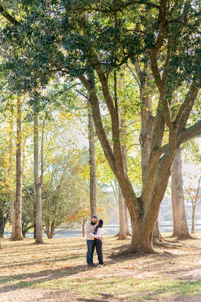 woman kisses man on the cheek with a tall tree next to them with fallen leaves on the ground during Greenville engagement session 