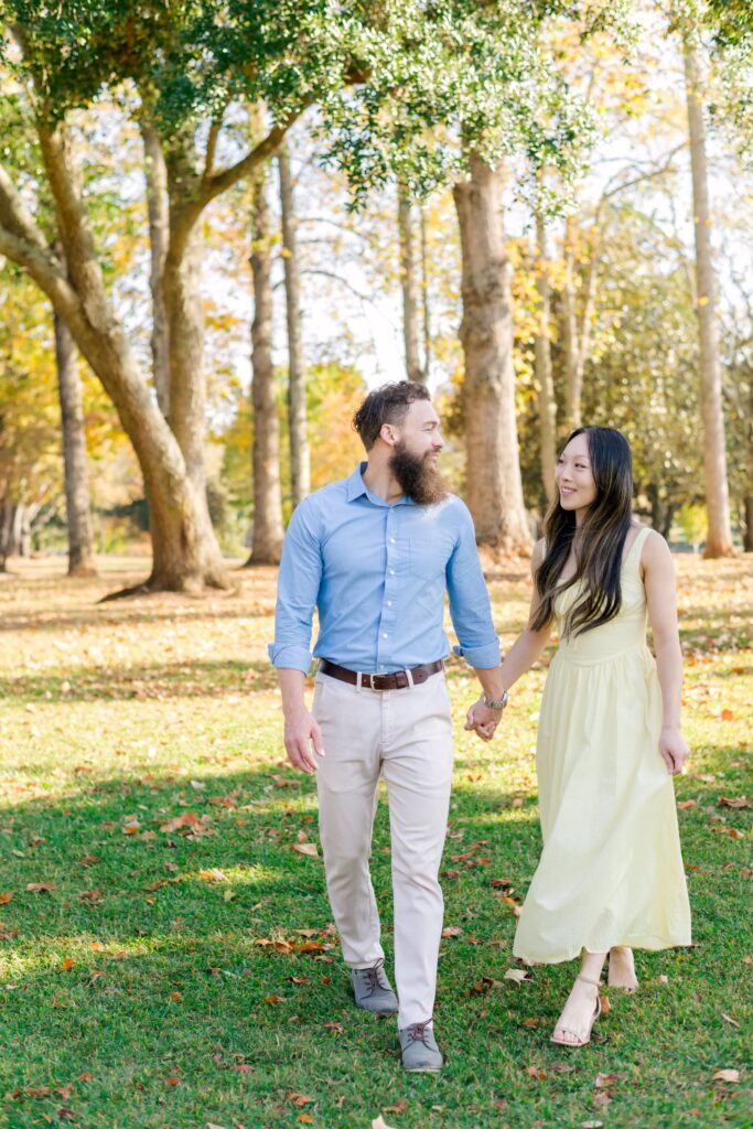 Man and woman walk in a field of fallen leaves looking at each other and smiling. Woman wears a yellow dress and man wears a blue button up shirt with khakis during Greenville engagement session 