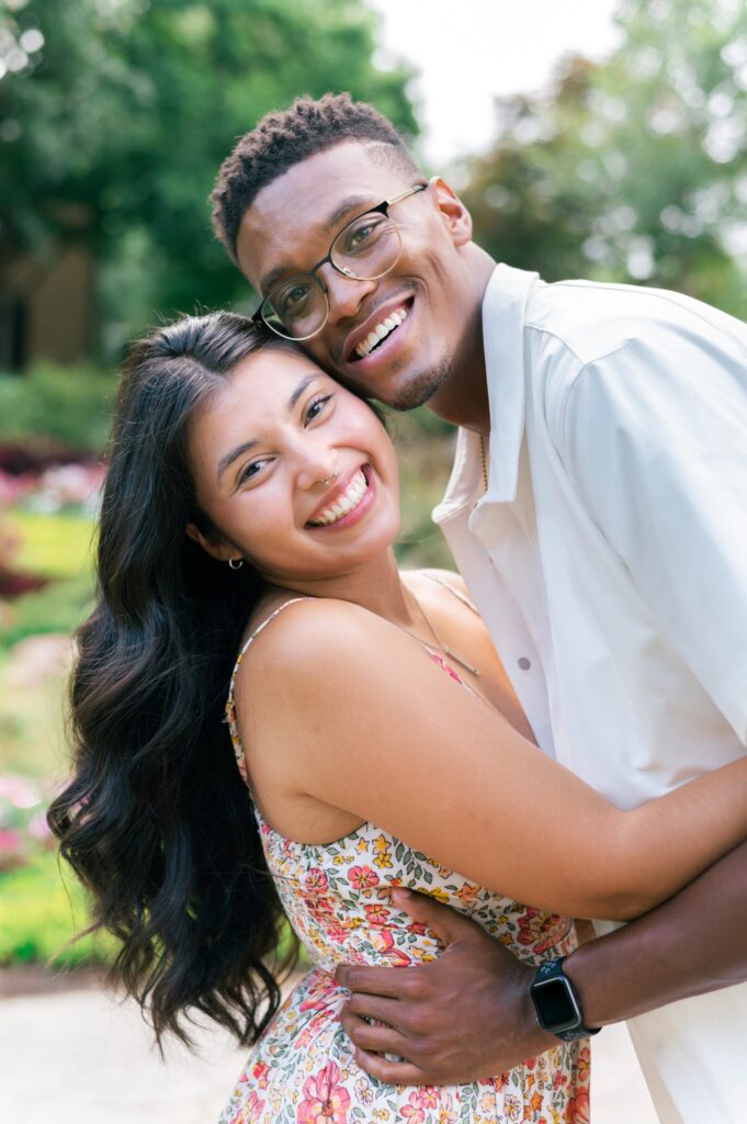 woman and man embrace in front of a field of flowers on a stone path looking at the camera and smiling during Greenville engagement session 