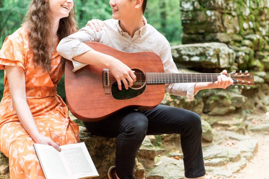 man has a guitar playing for his fiancé while she holds a book and smiles while they sit on rocks during a Greenville engagement session 