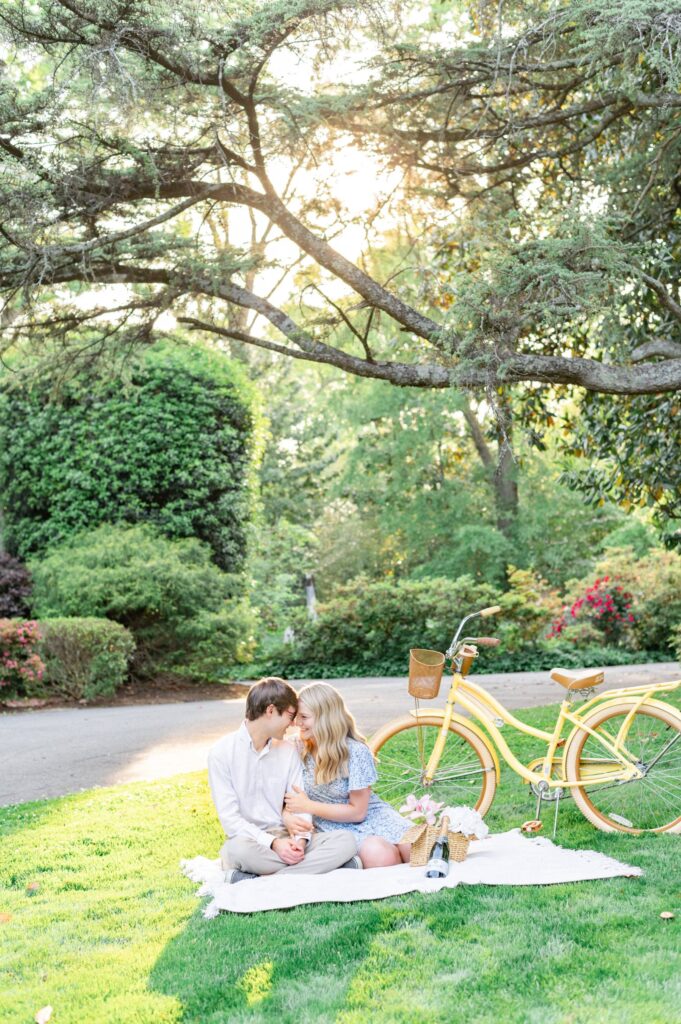 man and woman sit on a picnic blanket with a yellow bike behind them with a pavement path behind them holding hands looking at each other during Greenville engagement session 