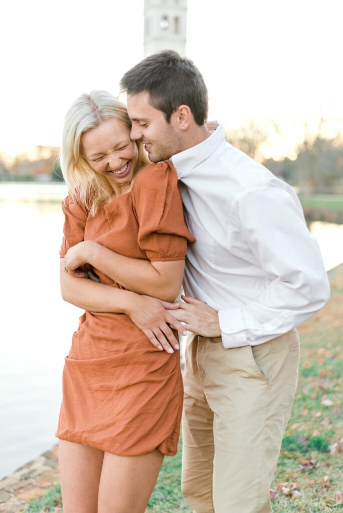 man holds his future wife whispering in her ear as she giggles in front of the water at Furman University engagement session