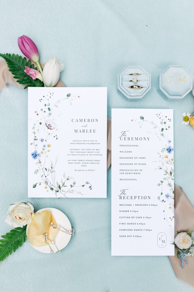 detail shots of save the dates/invitations with rings, boutonniere and flowers with a blue background taken at Piney Grove Ranch Wedding