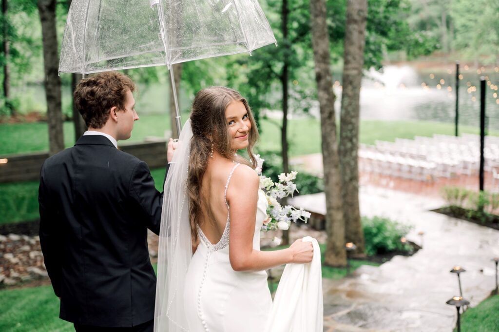 bride and groom walk away from camera while bride looks back holding an bouquet of flowers and groom holds umbrella at Piney Grove Ranch wedding