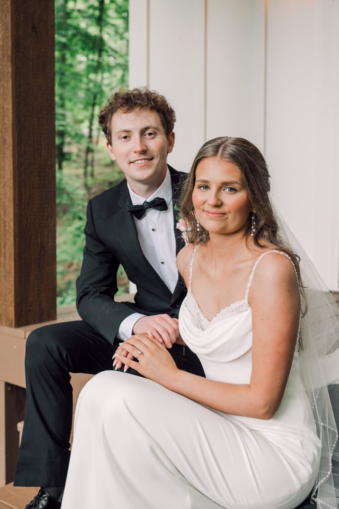 Bride and groom look at the camera sitting on stairs, the bride is one step below groom with her hand on groom's knee showing off her wedding ring at Piney Grove Ranch wedding