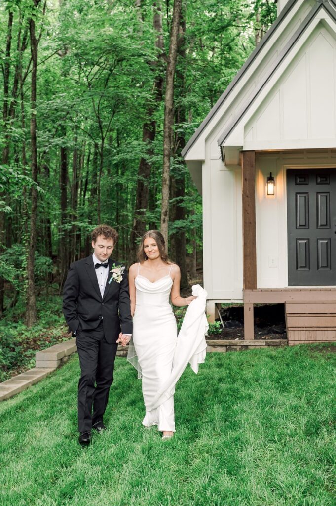 Bride looks at camera holding up the train of her dress while groom holds her hand and looks down at the grass as they're standing in front of a white building at Piney Grove Ranch wedding