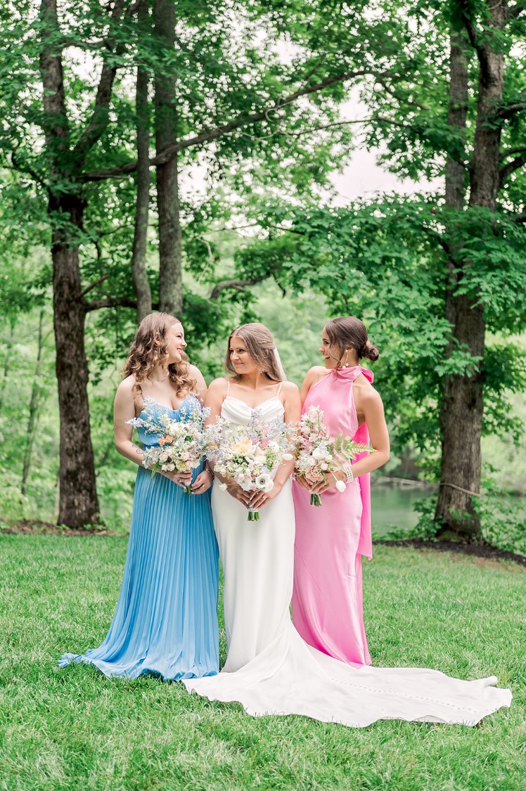 Bride stands with two bridesmaids one wearing pink dress the other wearing blue dress holding flowers at Piney Grove Ranch Wedding