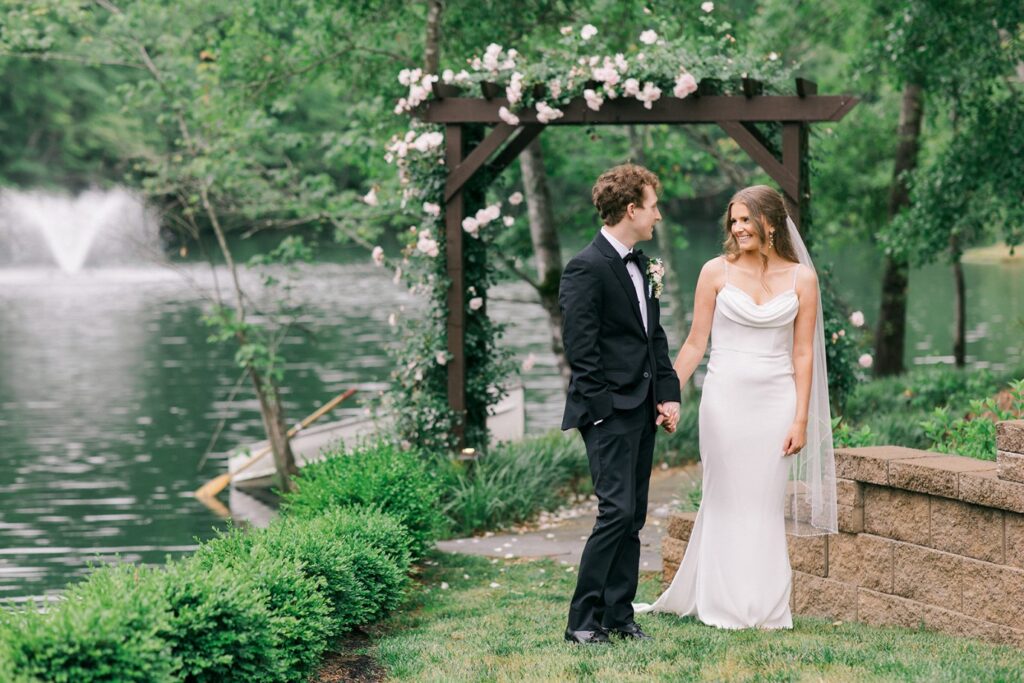Bride and groom look at each other smiling in front of an arch with pink flowers with a river and a canoe behind it at Piney Grove Ranch wedding