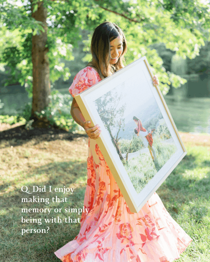 Female photographer in a pink flowy dress twirling with 1 framed photo outdoors in front of a tree and lake.