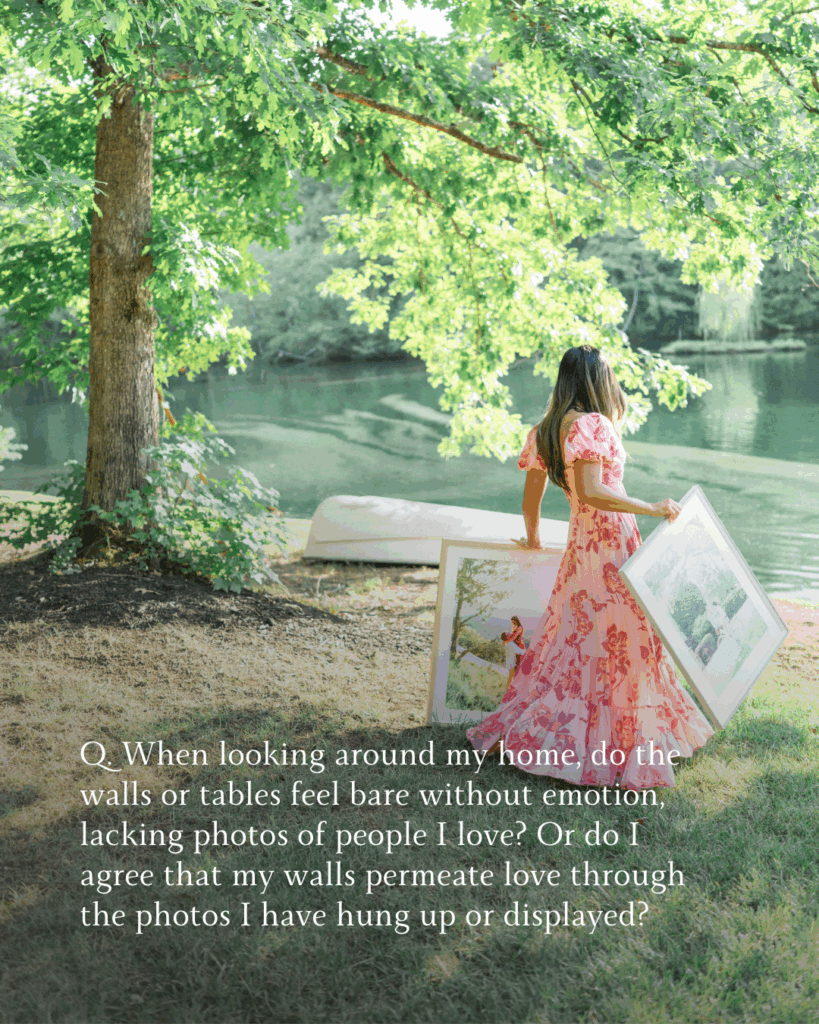 Female photographer in a pink flowy dress walking with two framed photos outdoors in front of a tree, upturned rowboat, and lake.