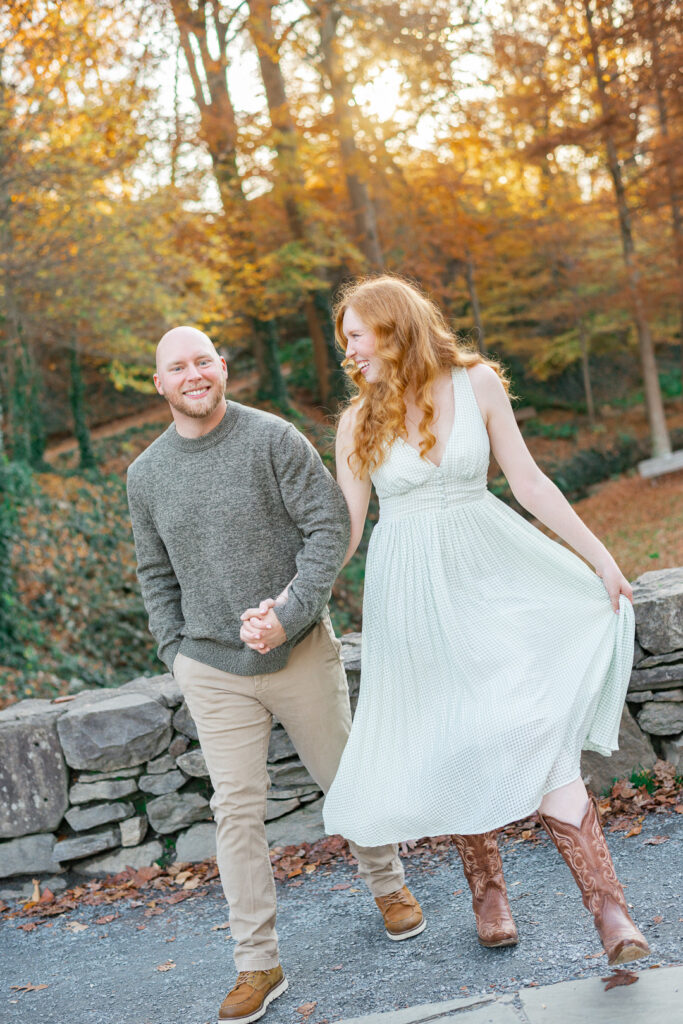 Couple holding hands and laughing while walking beside a stone wall in an autumn park, with colorful fall leaves in the background.