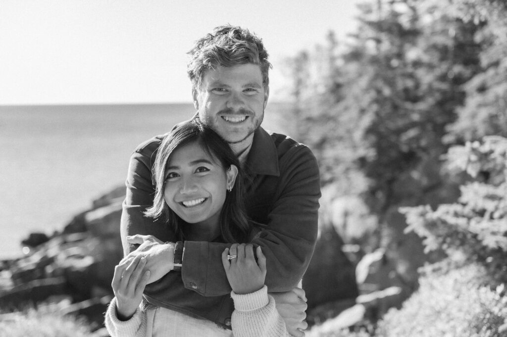 Black-and-white photo of Abby Joy Studio and her husband smiling as he hugs her from behind outdoors by the coast.