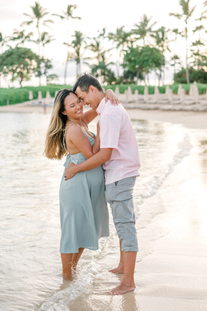 Couple hugging and smiling while standing barefoot in the ocean at sunset with palm trees in the background.