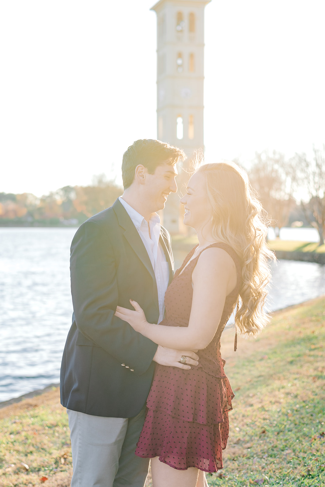 Newly engaged couple in a blazer and red dress laugh by a lake at sunset