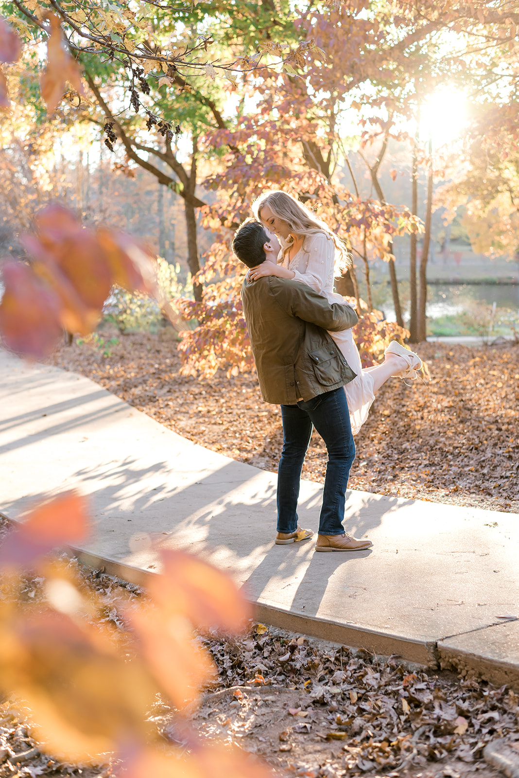 A man lifts his fiancee in a park sidewalk in fall at sunset for a kiss