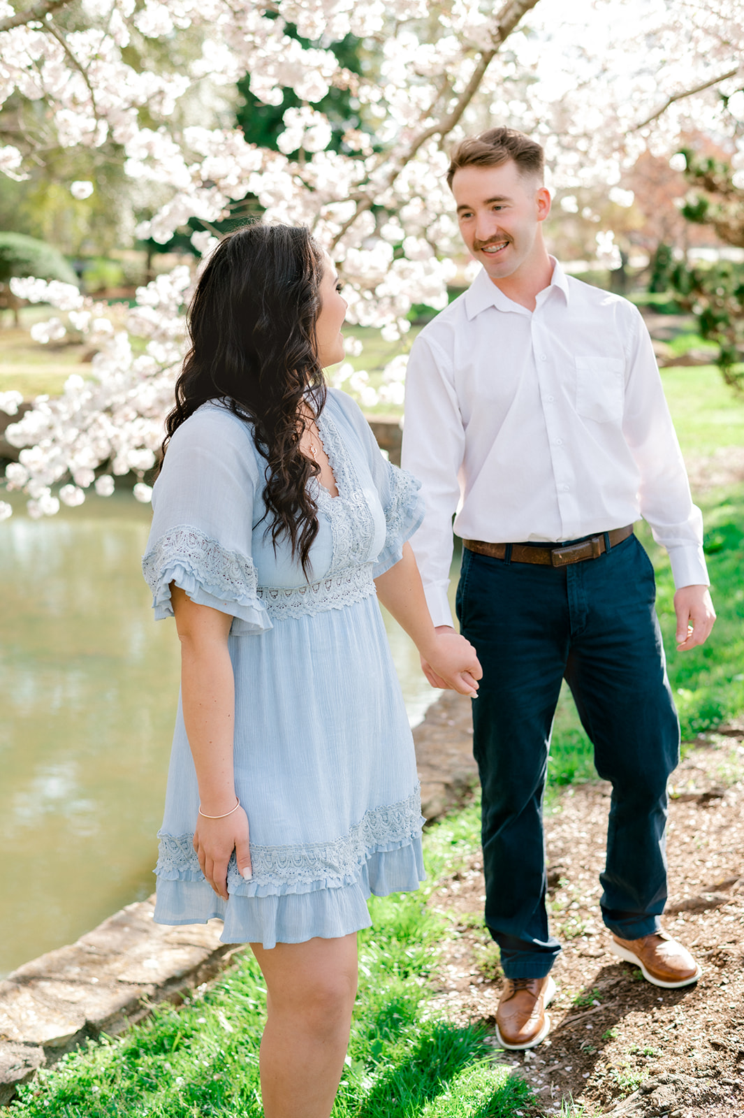 A happy engaged couple walk in a garden under a blooming tree holding hands and smiling at each other in a white shirt and blue dress by a pond after visiting romantic restaurants in Greenville, SC