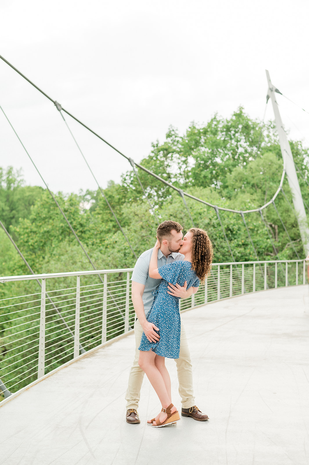 A newly engaged couple kisses on a pedestrian bridge in blue dress before heading to romantic restaurants in Greenville, SC