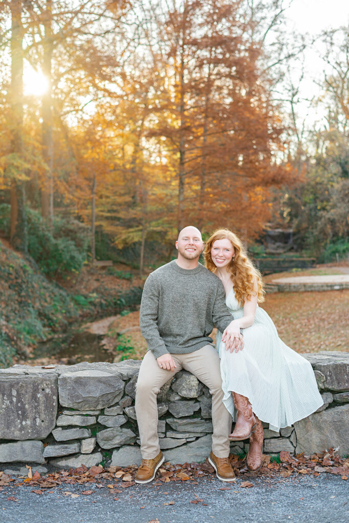 A man in a grey sweater sits on a stone wall at sunset with his fiancee in white in a park in fall before heading to romantic restaurants in Greenville, SC