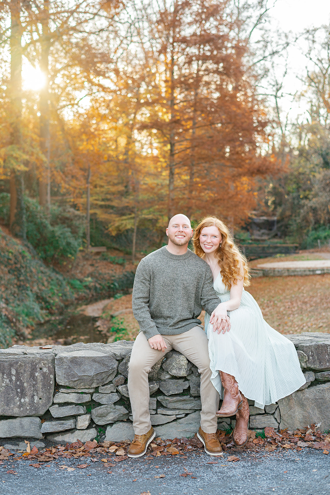 A man in a grey sweater sits on a stone wall at sunset with his fiancee in white in a park in fall before heading to romantic restaurants in Greenville, SC
